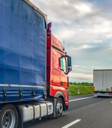 A red and blue freight truck drives along a highway beside another truck under a cloudy sky. Useful for logistics websites, supply chain articles, freight transport content, and industry presentations.